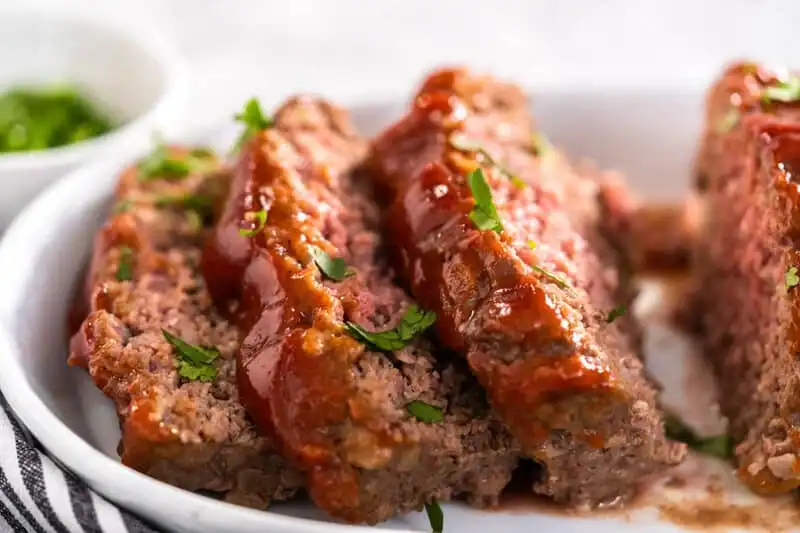 Three slices of meatloaf topped with ketchup and garnished with chopped parsley are served on a white plate.