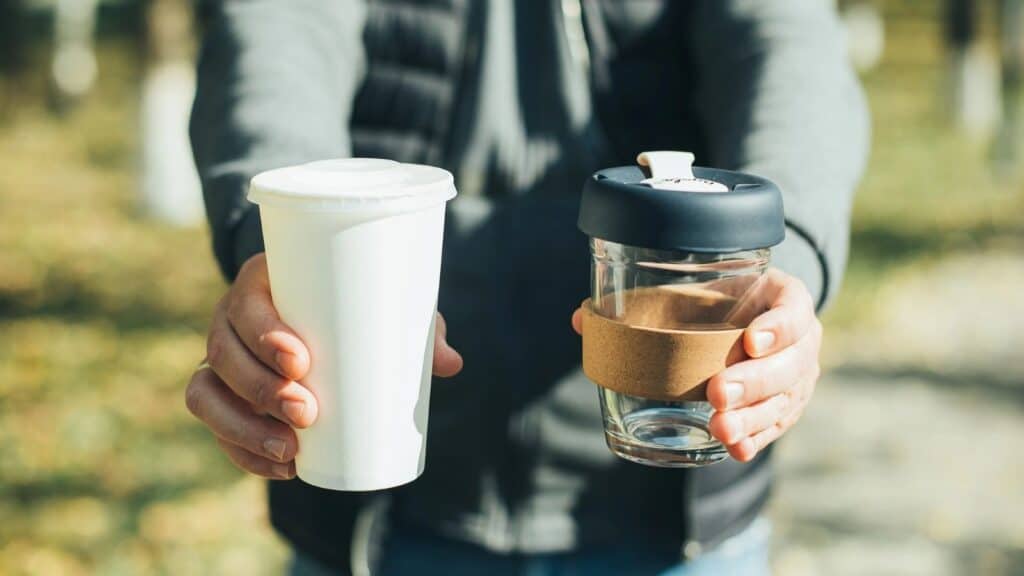 A person holds a disposable white paper cup in one hand and a reusable glass coffee cup with a black lid and cork band in the other hand.