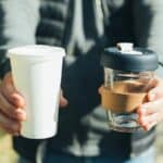 A person holds a disposable white paper cup in one hand and a reusable glass coffee cup with a black lid and cork band in the other hand.