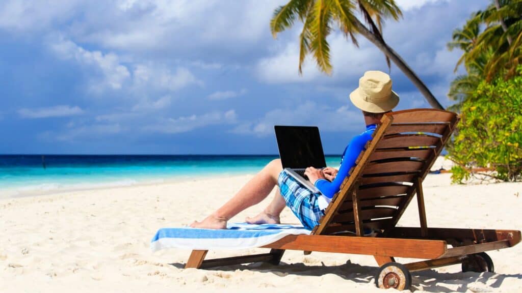Enjoying a bleisure travel moment, a person relaxes on a lounge chair on a sandy beach, using a laptop with palm trees and the ocean in the background under a partly cloudy sky.