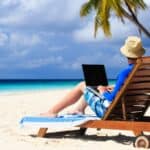 Enjoying a bleisure travel moment, a person relaxes on a lounge chair on a sandy beach, using a laptop with palm trees and the ocean in the background under a partly cloudy sky.