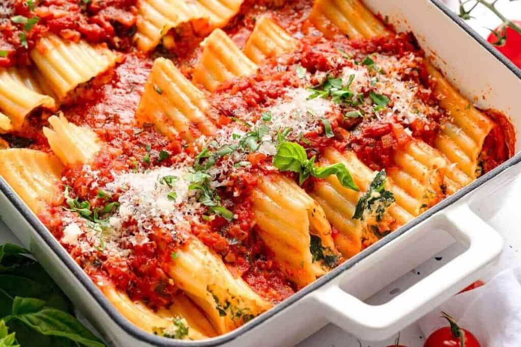 Baked manicotti pasta filled with ricotta and spinach, topped with tomato sauce, grated cheese, and fresh basil in a white casserole dish.