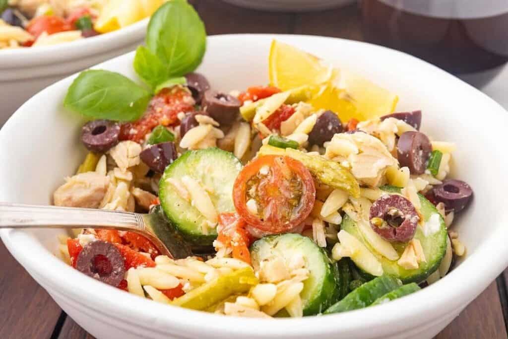 A bowl of orzo pasta salad with cucumbers, cherry tomatoes, olives, basil, and a lemon wedge, with a fork resting inside.