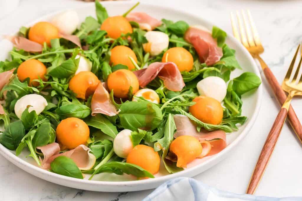 A plate of salad with arugula, cantaloupe balls, mozzarella balls, and slices of prosciutto, served on a white plate with gold and wooden utensils beside it.