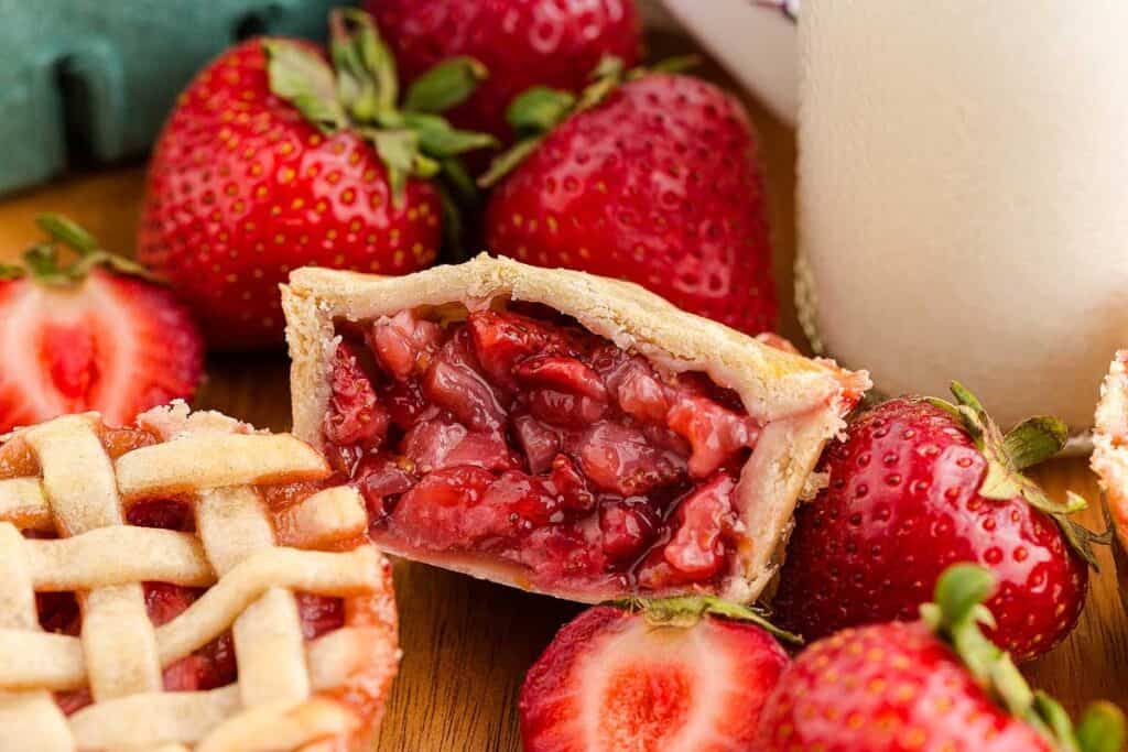 A close-up of a mini strawberry pie with a lattice crust, surrounded by fresh whole and sliced strawberries on a wooden surface.