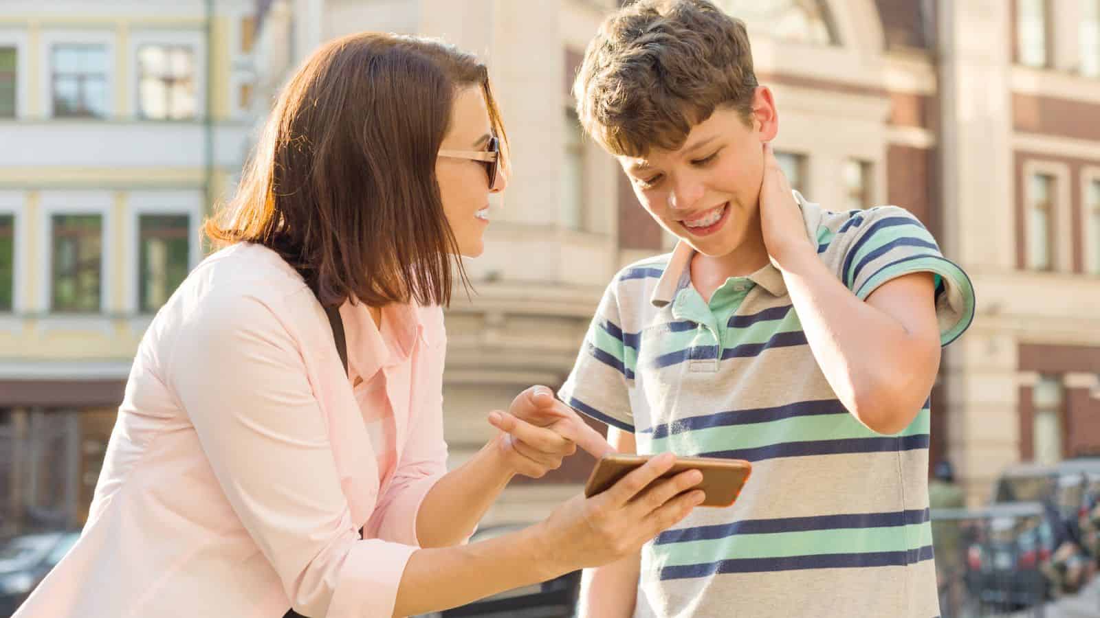 A woman shows a smartphone to a smiling teenage boy outdoors; they are engaged in conversation near buildings.