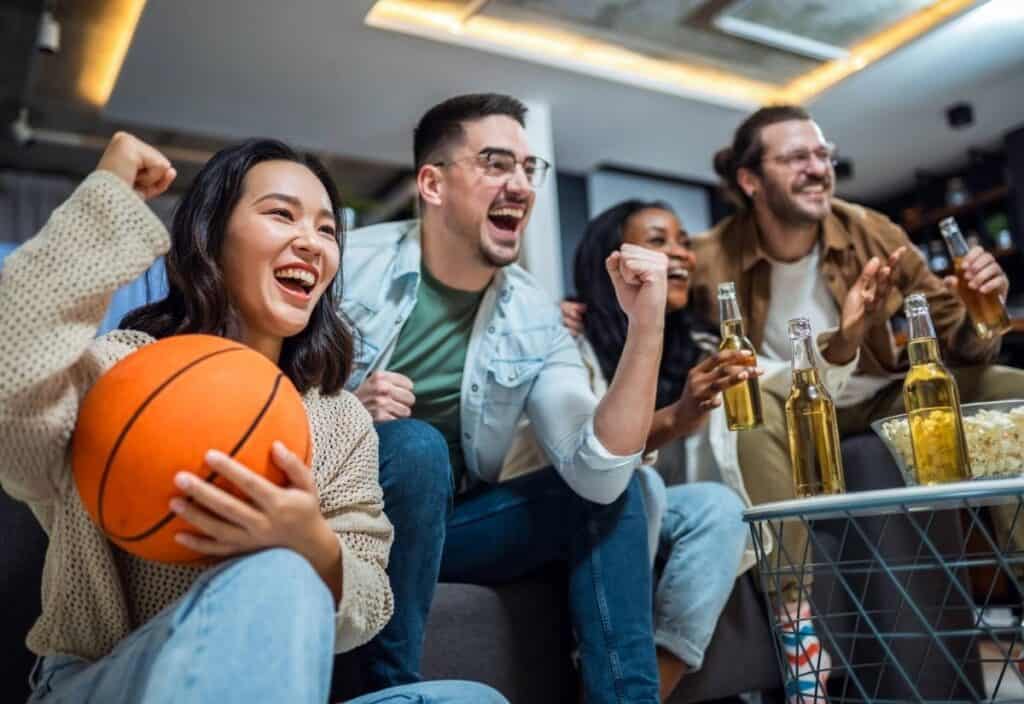Four people sit on a couch cheering while watching a game on TV, with drinks, snacks, and a basketball in the foreground.