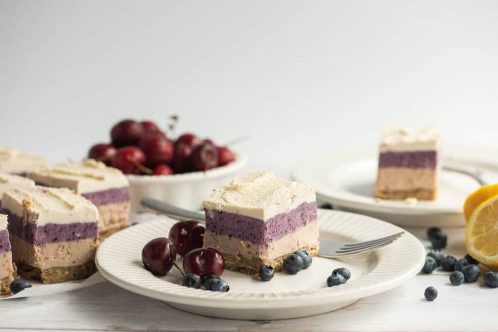 A plate with a layered slice of cheesecake garnished with blueberries and cherries, with more cheesecake slices, fruit, and a lemon visible in the background.