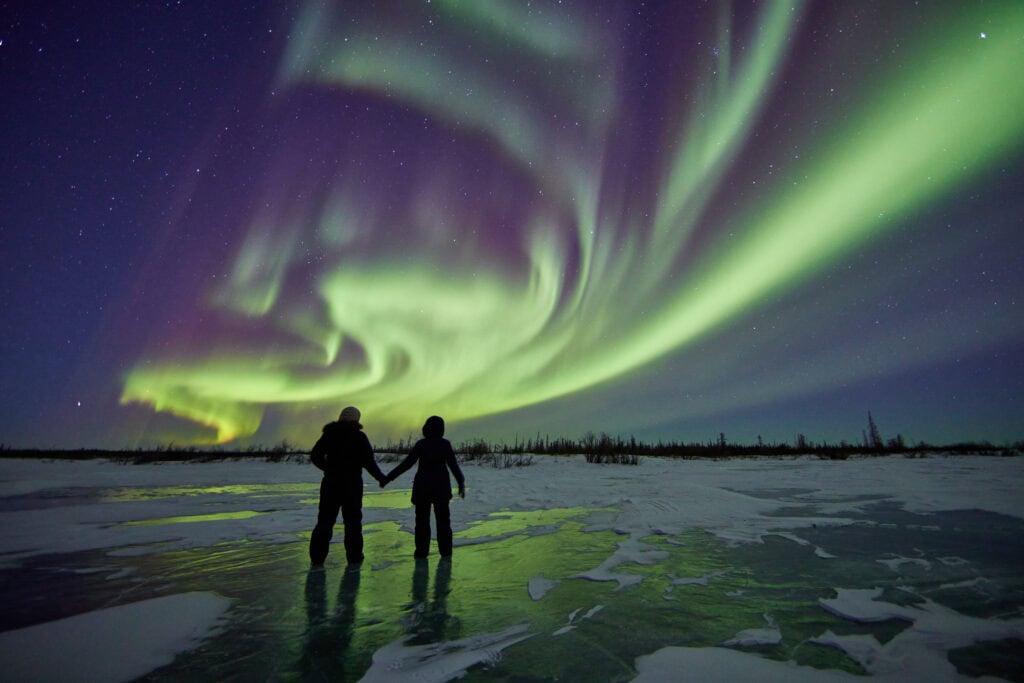 Two people stand on a snowy, frozen landscape at night, watching green and purple aurora borealis lights in the sky.