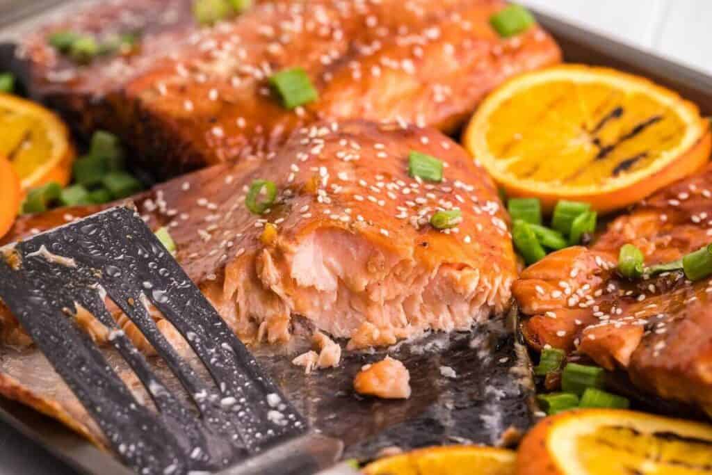Baked salmon fillet with a flaky texture, garnished with sesame seeds and chopped green onions, surrounded by orange slices on a tray with a metal spatula.