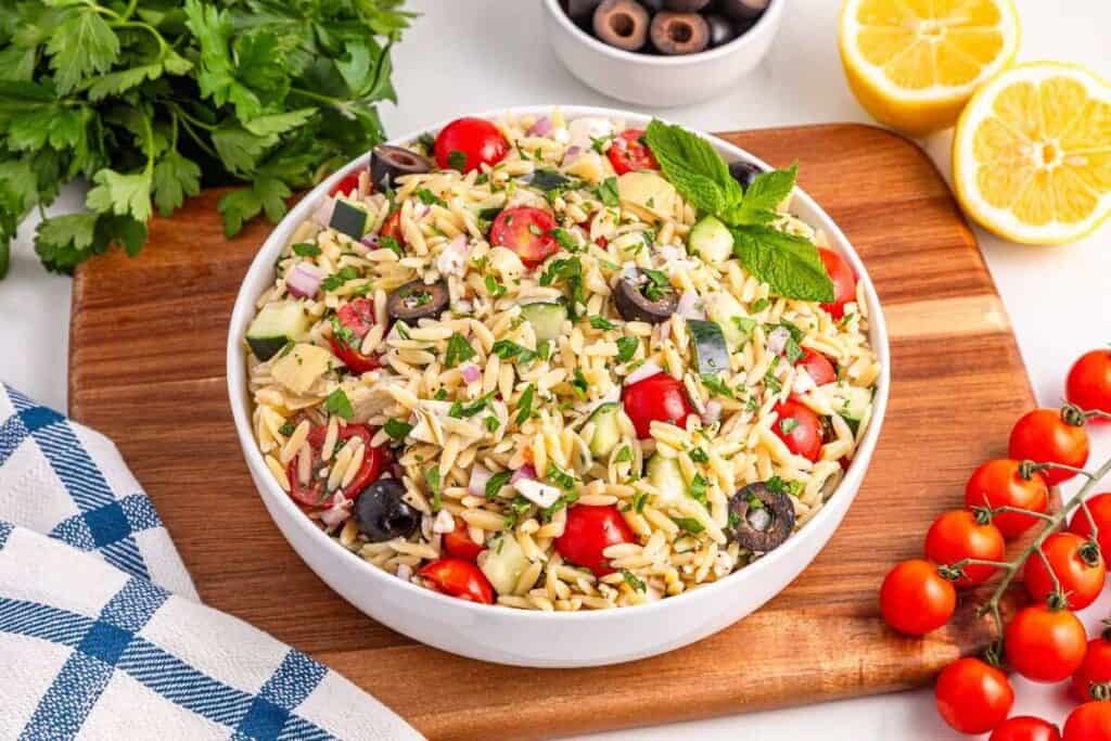 A bowl of orzo pasta salad with cherry tomatoes, cucumbers, black olives, red onions, and herbs, placed on a wooden board with fresh parsley, lemons, and tomatoes nearby.