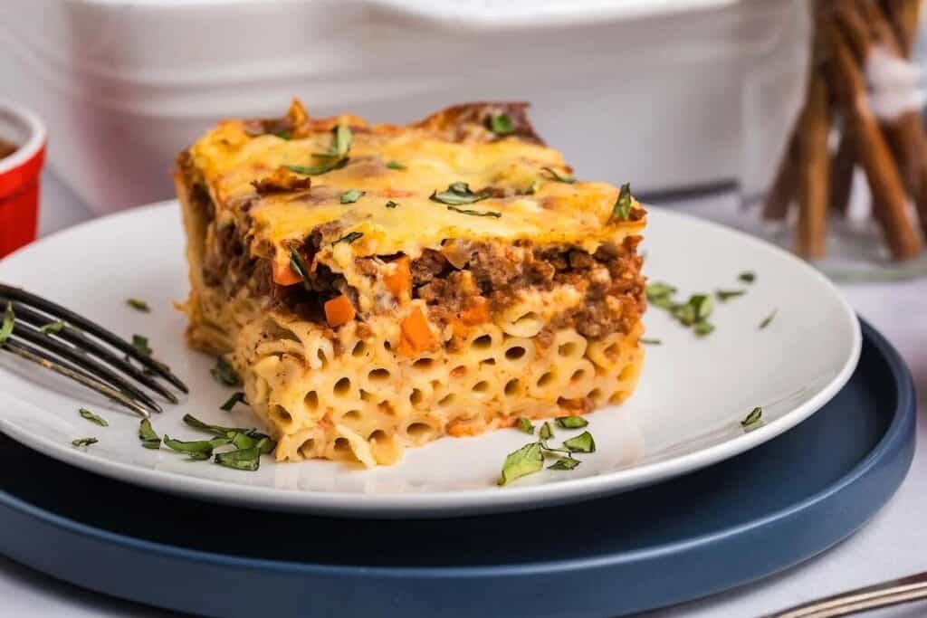 A square slice of baked pasta with ground meat and cheese sits on a white plate, garnished with chopped herbs, next to a fork and knife.