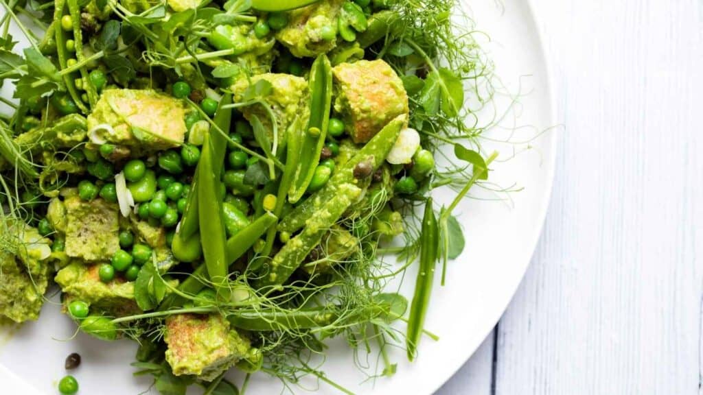 A plate of fresh green salad with avocado, snap peas, peas, microgreens, and herbs on a white background.