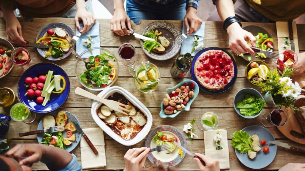 People eating a meal together at a wooden table, with various dishes, salads, vegetables, bread, and drinks spread across the surface.