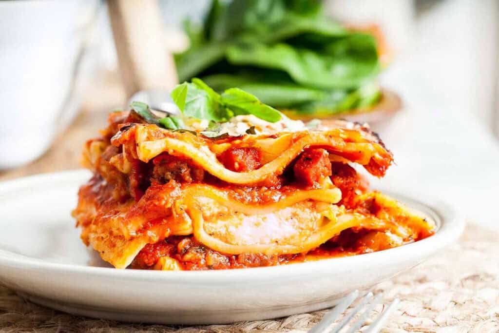 A slice of lasagna with layers of pasta, meat sauce, and cheese is served on a white plate, garnished with fresh basil.