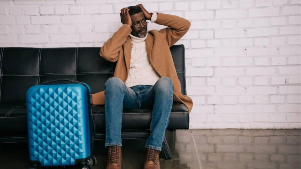 A man sits on a black couch next to a blue suitcase, holding his head with both hands and looking distressed&mdash;perhaps wishing he had travel insurance.