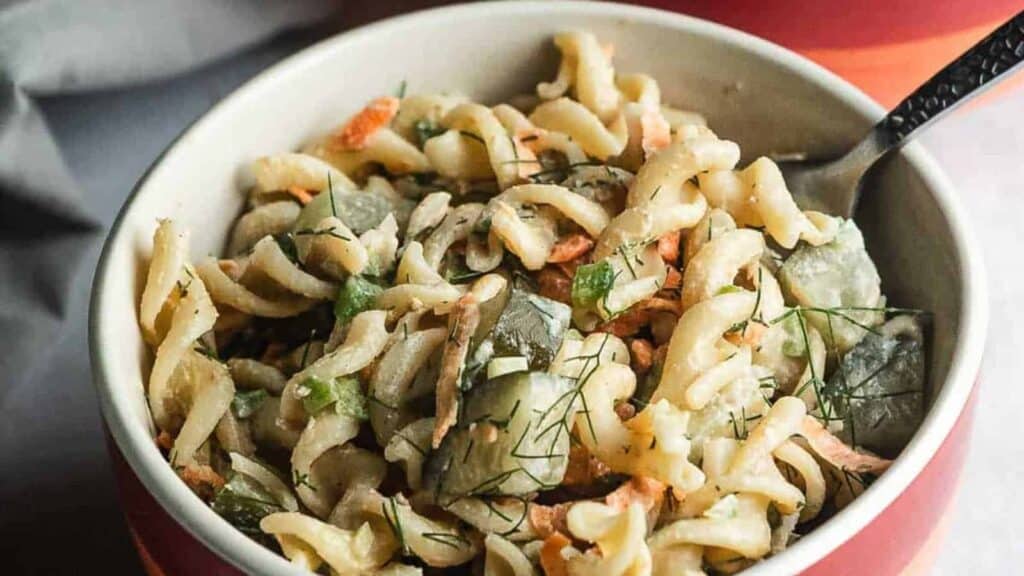 A bowl of rotini pasta salad with chopped cucumbers, grated carrots, and fresh herbs, served with a fork.