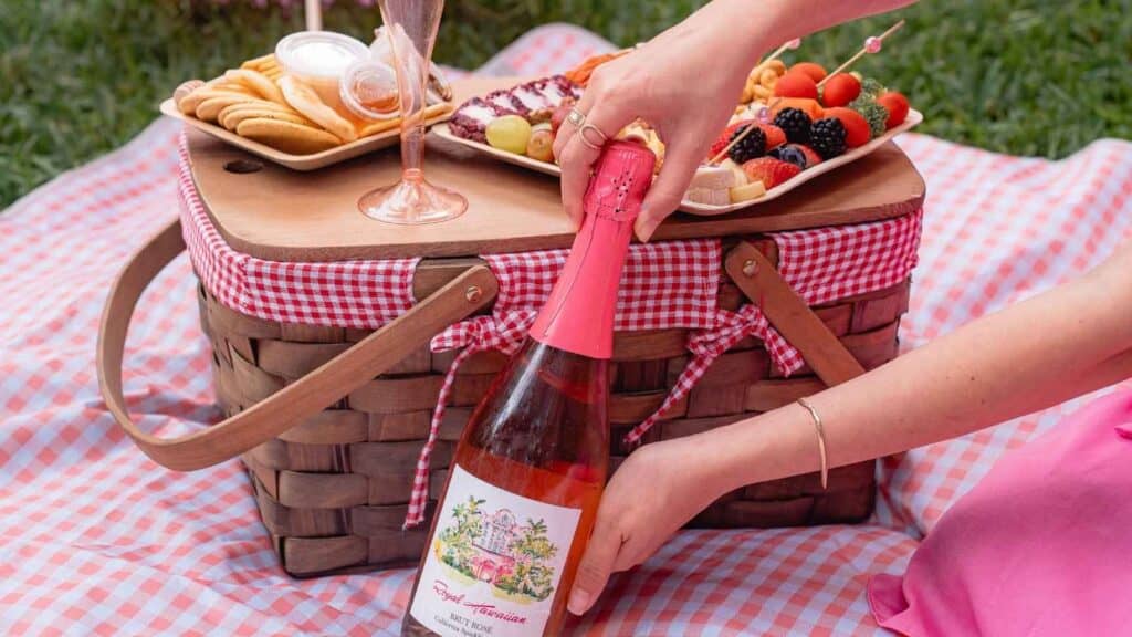 A hand opens a bottle of ros&eacute; wine on a picnic basket with a gingham cloth. A platter of crackers, fruit, and cheese sits on top of the basket.