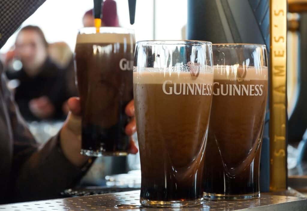 Two pints of Guinness sit on a bar counter, with another glass of the famous stout being handed to someone in the background.