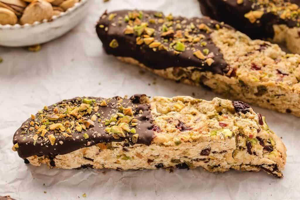 Two biscotti cookies partially dipped in chocolate and sprinkled with chopped pistachios rest on parchment paper, with a bowl of unshelled pistachios in the background.