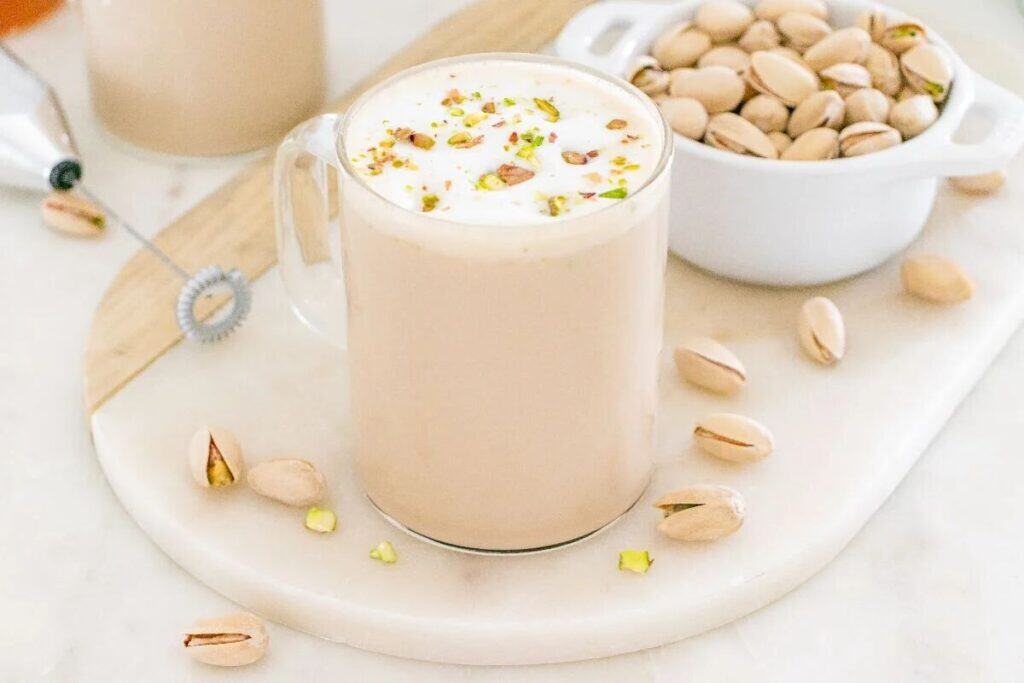 A glass mug of pistachio latte topped with chopped pistachios, next to a white bowl of shelled pistachios and scattered pistachios on a white serving board.