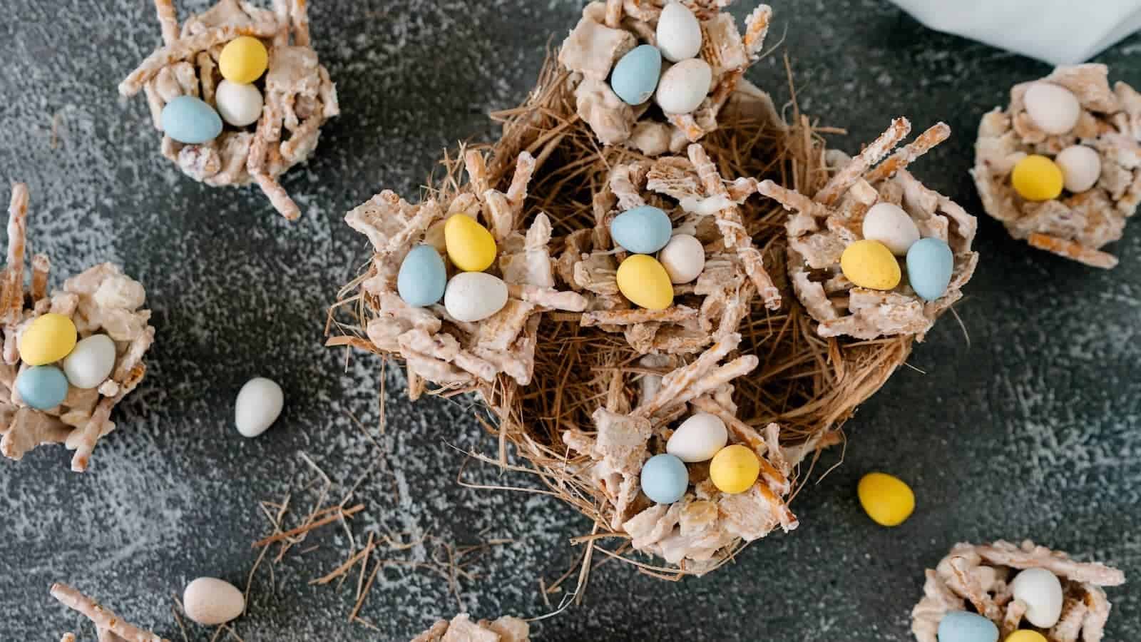 A close-up of a homemade edible bird nest treat made from cereal sticks and white coating, filled with candy eggs, on a textured surface.