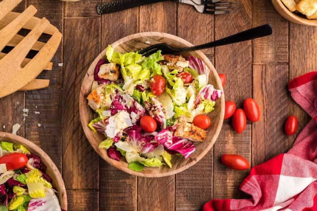 A wooden bowl of salad with lettuce, cherry tomatoes, croutons, and shredded cheese, placed on a wooden table beside a red checkered napkin and wooden utensils.