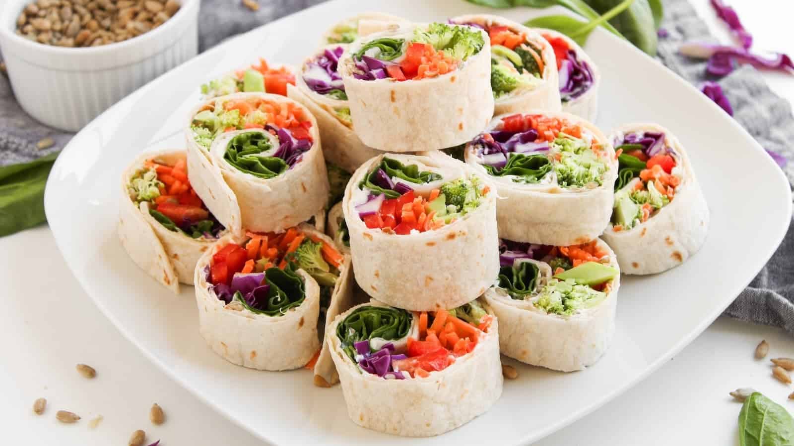 A white plate with neatly stacked veggie pinwheel wraps filled with colorful vegetables, next to a small bowl of seeds and scattered spinach leaves.