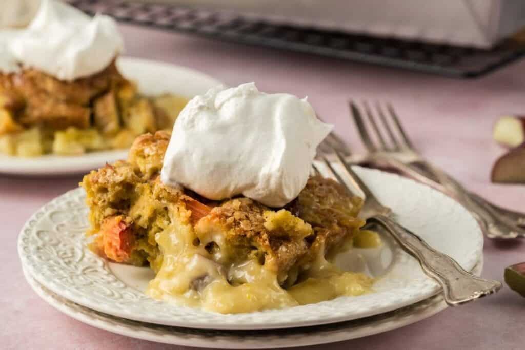 A slice of rhubarb pie with a golden crust and creamy filling, topped with whipped cream, served on a white plate with two forks.