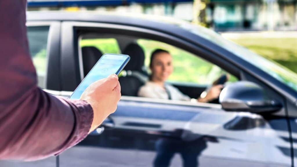 Person holding a smartphone stands outside while a driver in a car looks out of the window and smiles, suggesting a rideshare or pickup situation.
