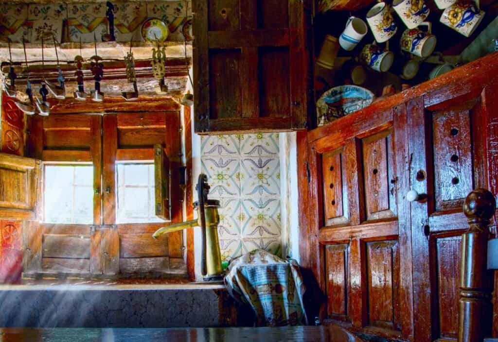 A rustic kitchen with wooden cabinets, a brass water pump, hanging mugs, utensils above a window, and decorative patterned tiles&mdash;perfect for cooking like grandmothers did.