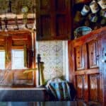 A rustic kitchen with wooden cabinets, a brass water pump, hanging mugs, utensils above a window, and decorative patterned tiles—perfect for cooking like grandmothers did.