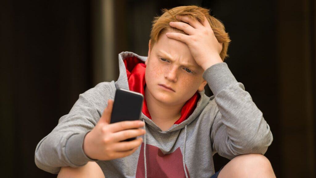 A boy with red hair wearing a gray hoodie looks at his phone with a concerned expression, holding his head with one hand.