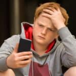 A boy with red hair wearing a gray hoodie looks at his phone with a concerned expression, holding his head with one hand.