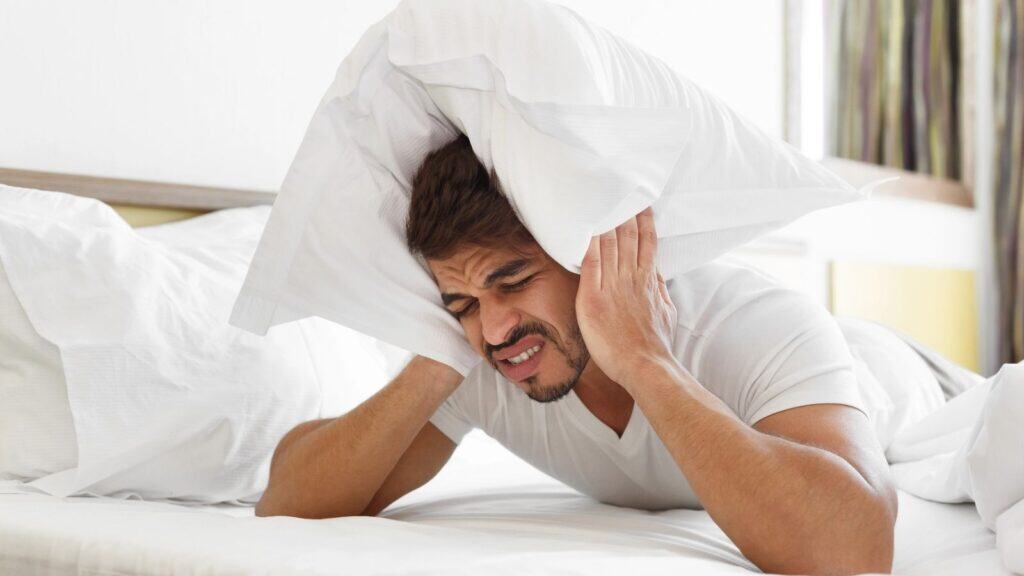 A man in a white shirt lies in bed covering his ears with a pillow, appearing frustrated or disturbed by noise.