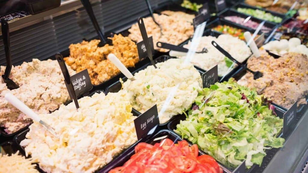 A deli counter displays various prepared salads including egg salad, tuna salad, greens, tomatoes, and other mixed dishes in black trays with serving spoons.
