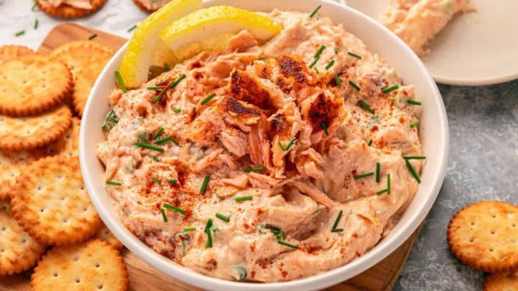 A bowl of salmon dip garnished with lemon slices, chives, paprika, and flaked salmon, surrounded by round crackers on a gray surface.