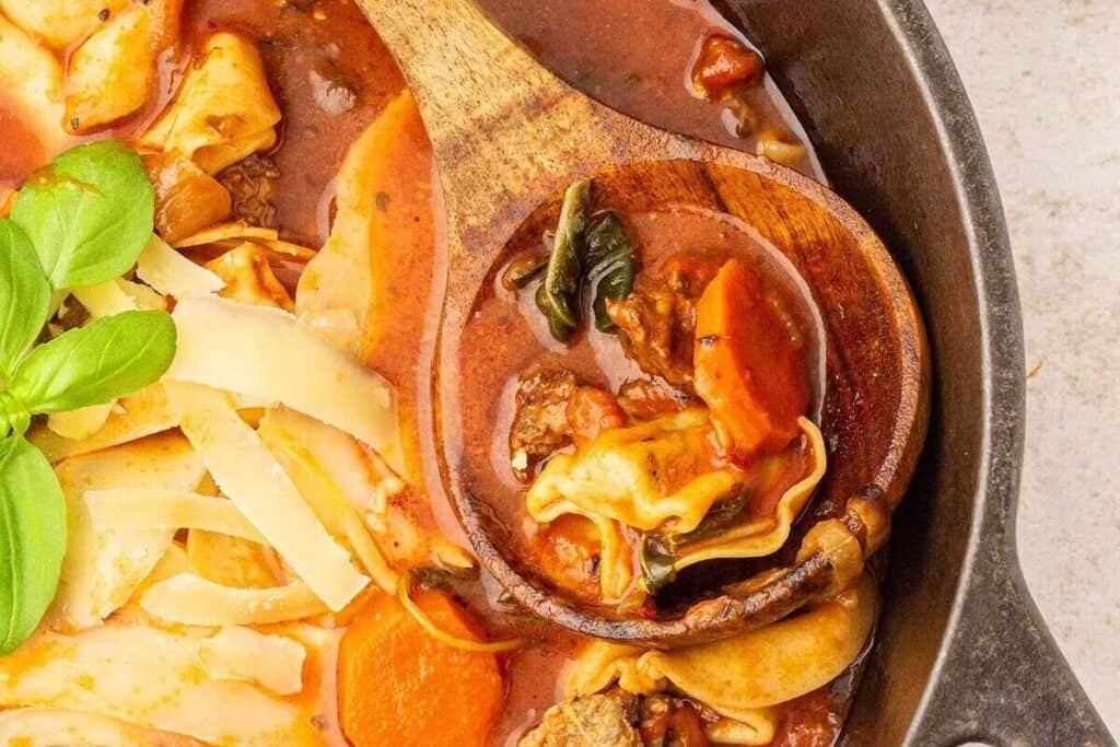 A wooden spoon rests in a pot of lasagna soup with visible pasta, carrots, spinach, tomato broth, beef, and shredded cheese.