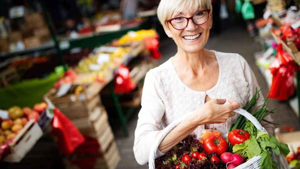 Older woman with short gray hair and glasses holding a basket of fresh vegetables at a fresh farmers market, smiling at the camera.