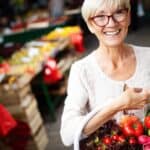 Older woman with short gray hair and glasses holding a basket of fresh vegetables at a fresh farmers market, smiling at the camera.
