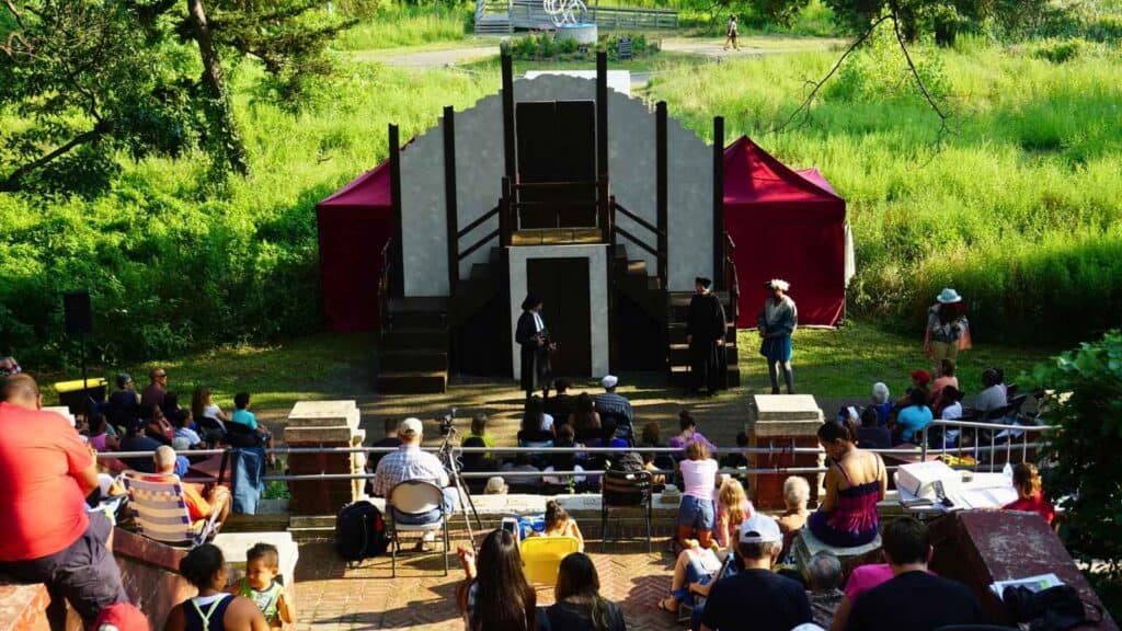 Outdoor theater performance with actors on stage and a seated audience watching, surrounded by greenery and natural light.
