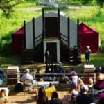 Outdoor theater performance with actors on stage and a seated audience watching, surrounded by greenery and natural light.
