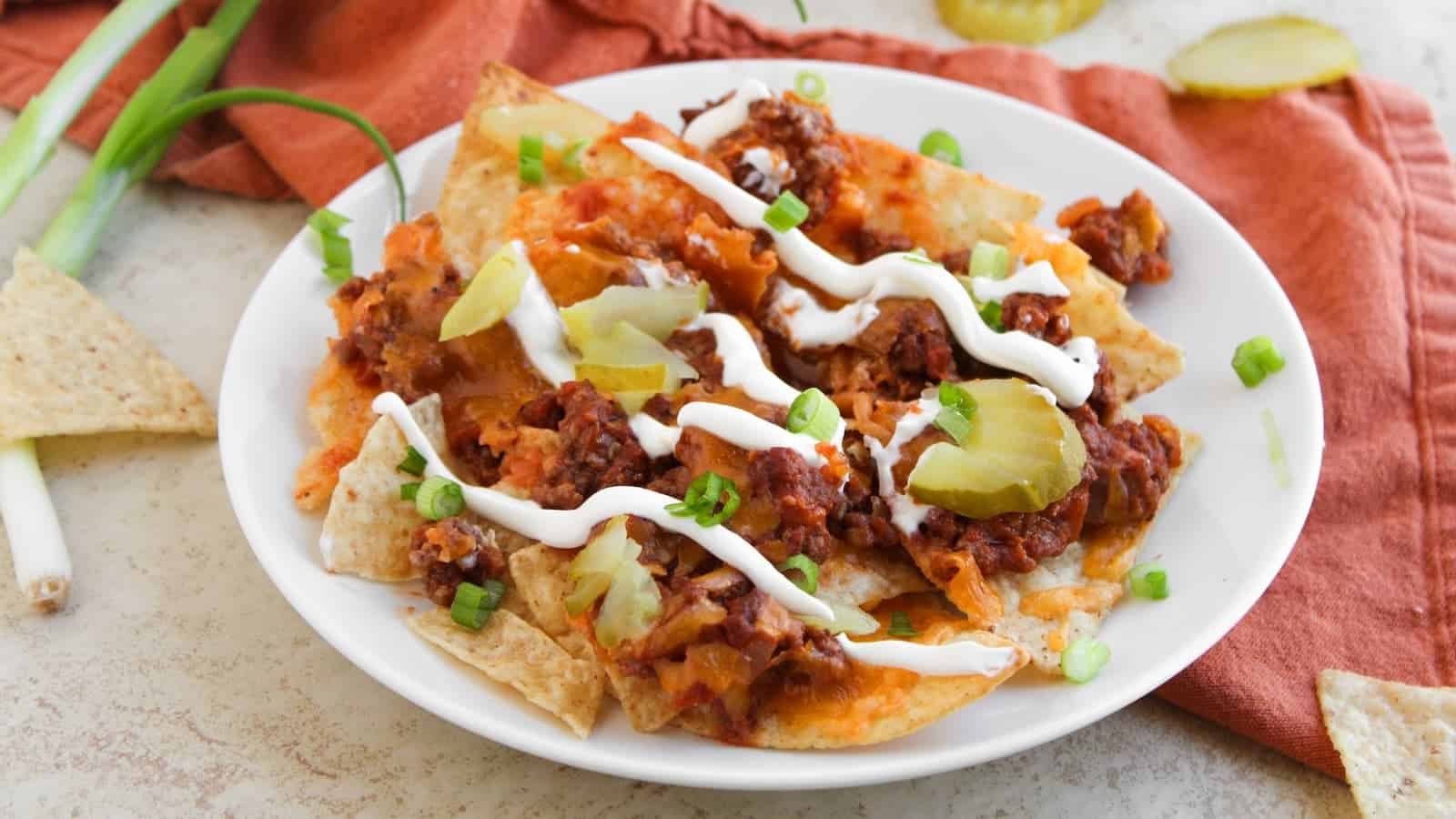 A plate of nachos topped with ground beef, melted cheese, pickle slices, sour cream, and chopped green onions, with tortilla chips and green onions beside the plate.