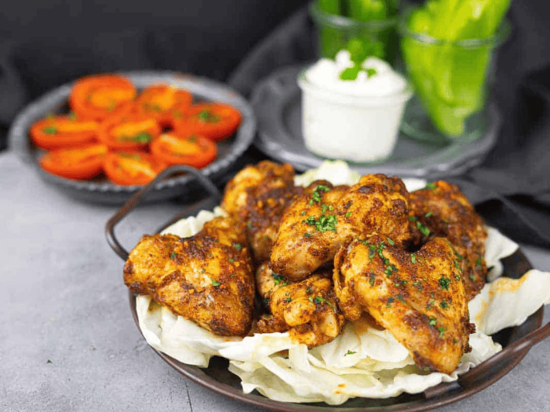 A plate of seasoned grilled chicken wings on a bed of cabbage, with sliced tomatoes, lettuce leaves, and a bowl of dipping sauce in the background.