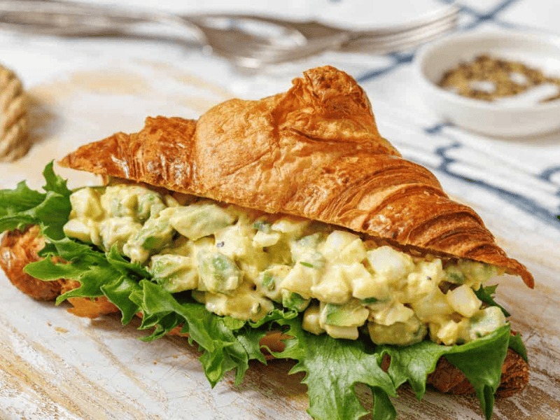 A croissant sandwich filled with Avocado egg salad, lettuce, and herbs sits on a wooden board.