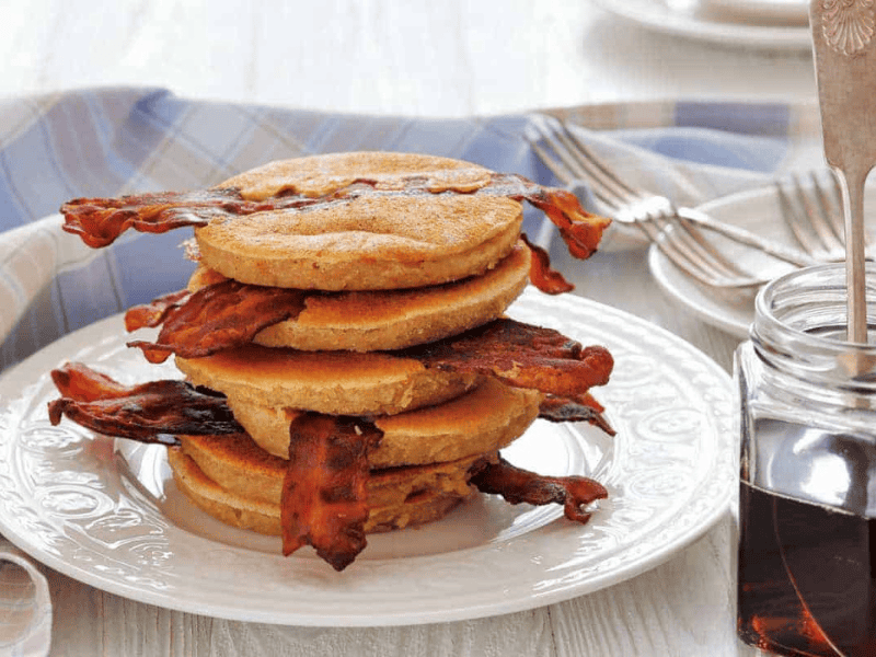 A stack of pancakes with strips of bacon between layers sits on a white plate, next to a jar of syrup and a folded napkin with forks.