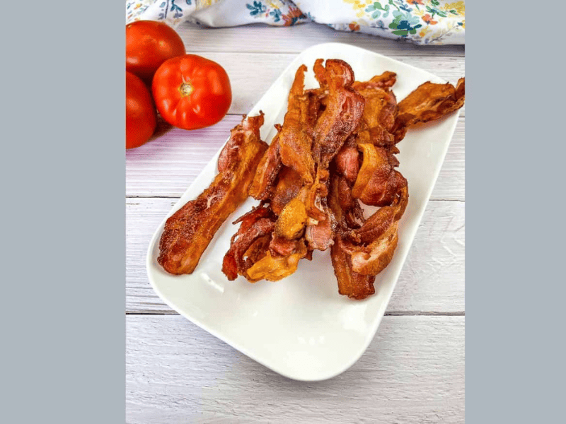 Bacon strips on a white plate next to tomatoes.
