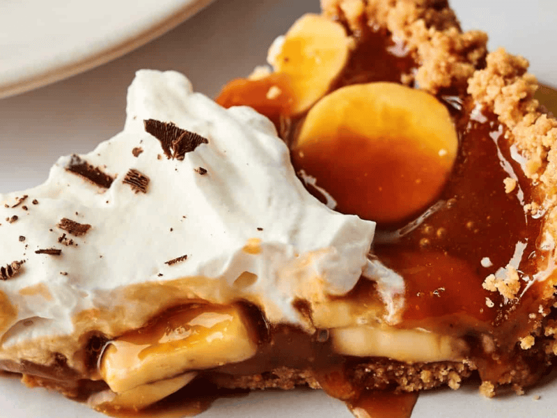 A slice of banoffee pie with layers of banana, caramel, whipped cream, and a crumbly biscuit crust on a white plate.