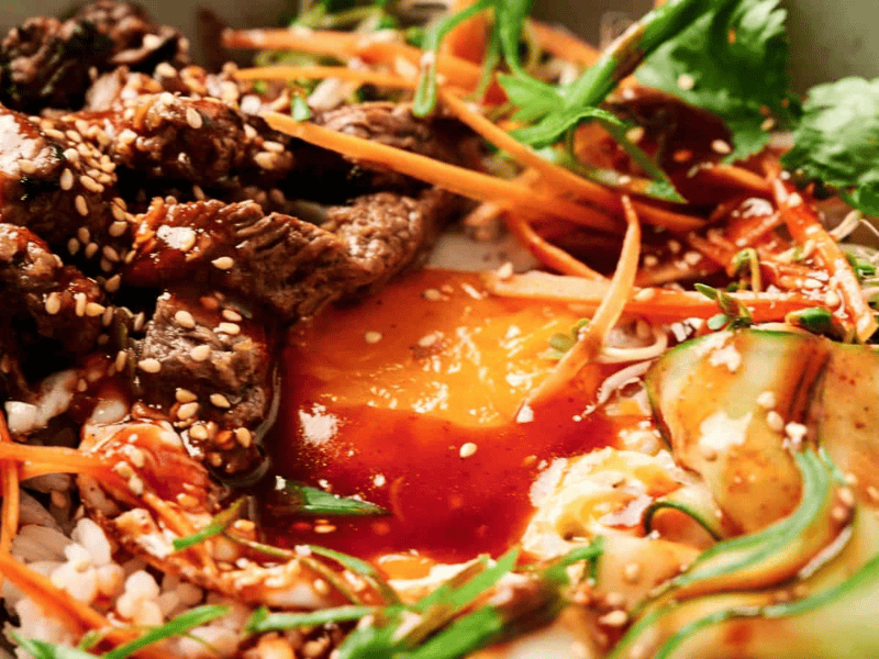 A bowl of bibimbap with sliced beef, egg yolk, carrots, cucumber, greens, and bean sprouts, topped with sesame seeds and red sauce.