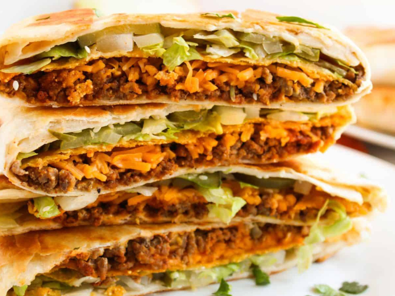 Close-up of a stack of folded tortillas filled with beef, melted cheese, lettuce, and onions on a white plate.